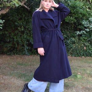 May include: A long, dark navy blue wool-blend coat featuring a wide lapel collar, long sleeves with folded cuffs, and a matching fabric tie belt at the waist. The coat is worn over light blue wide-leg denim jeans and black lace-up leather boots.