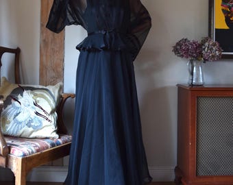 May include: A long black chiffon gown with a fitted bodice and a flowing skirt. The gown has a wide, ruffled neckline and a black satin sash at the waist.