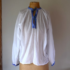 Op de afbeelding: Een wit, lang mouwig shirt met een blauwe en paarse geborduurde halslijn. Het shirt is op een houten mannequin.