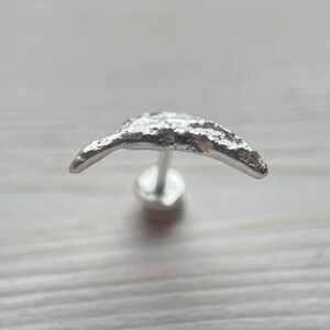 May include: A silver-colored stud earring with a crescent moon design. The earring has a textured surface and a post for insertion. The earring is set against a light-colored wooden background.