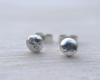 May include: Close-up of two small, silver stud earrings with a textured, hammered appearance. The earrings are round and slightly irregular in shape. The background is a light, neutral colour.
