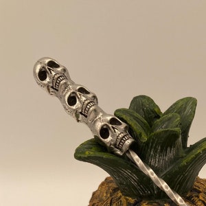 May include: A silver cocktail stirrer with three skull heads at the top, resting on a gold pineapple skull.