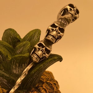 May include: A silver cocktail stirrer with a series of skull heads attached to the top. The stirrer is placed in a gold pineapple-shaped skull.