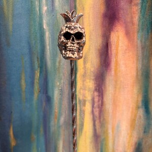 May include: A silver-colored bar spoon with a skull-shaped pineapple design. The skull has hollow eye sockets and a detailed jawline. The stem is twisted, and the background is a colorful abstract painting.