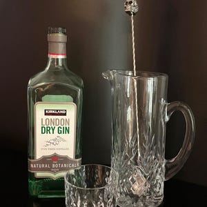 May include: A still life arrangement featuring a bottle of Kirkland London Dry Gin, a crystal pitcher, a rocks glass, and a bar spoon with a skull-shaped top. The gin bottle has a green label with white text. The glassware is clear.