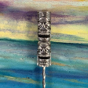 May include: A silver-colored bar spoon with a Tiki-style design. The Tiki head is carved with detailed facial features and stacked on top of each other. The spoon has a twisted handle.