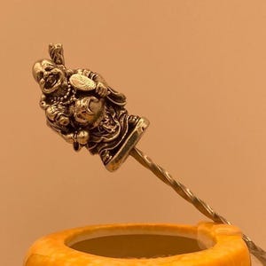 May include: A silver stir stick with a detailed figurine of a smiling Buddha holding a ball. The stir stick is resting on a yellow ceramic mug.