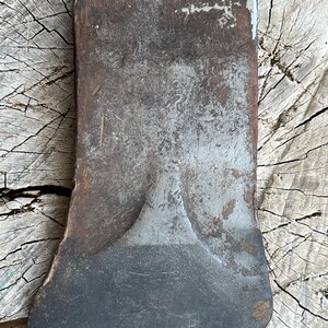 May include: A metal axe head featuring a weathered, dark grey finish with visible signs of oxidation and surface wear. The tool has a wide, curved cutting edge and a flat, rectangular poll, showcasing a textured, industrial appearance throughout.