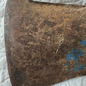 May include: A rusted metal axe head featuring a stamped logo with the letters HB inside a circle and the text MADE IN SWEDEN. The surface shows significant oxidation and patches of blue paint residue on the right side of the blade.