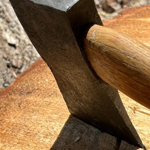Könnte beinhalten: Nahaufnahme eines Axtkopfes aus Metall mit Holzgriff, der auf einer Holzoberfläche liegt. Der Axtkopf ist dunkelgrau und der Griff hellbraun. Die Axt ist schräg positioniert, wobei die Klinge in das Holz eingelassen ist.
