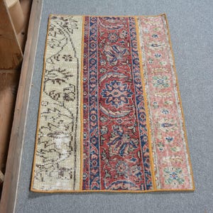 May include: Rectangular patchwork rug with three distinct sections. The left section is beige with a dark floral pattern. The centre is red with blue floral designs. The right section is pink with a floral pattern. The rug has a yellow border.