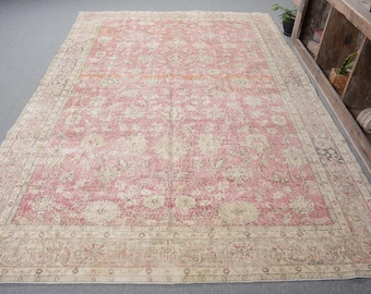 May include: A large rectangular area rug featuring a faded rose pink central field adorned with intricate cream and beige floral patterns. The rug is framed by a wide, detailed border in muted earth tones, showcasing repeating botanical motifs.