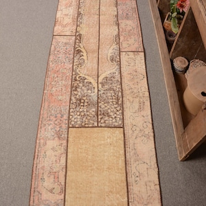 Pode incluir: Um tapete comprido e retangular de patchwork com fundo bege e detalhes em marrom, rosa e azul. O tapete é feito de várias peças de tecido diferentes costuradas juntas.