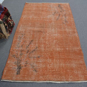 May include: An orange-toned, rectangular rug with a distressed appearance. The rug features a faded floral design in dark gray. The rug's edges are frayed, and it is placed on a gray surface. A wooden tool and a patterned bag are visible in the background.
