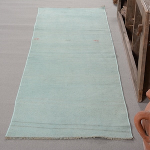 May include: A rectangular runner rug in a solid light mint green color. The rug features a flat-woven texture with subtle horizontal lines and small, sparse red embroidered accents. The short ends of the rug are finished with a short, light-colored fringe.