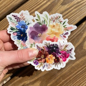 Op de afbeelding: Twee aquarelbloemenstickers met een witte achtergrond. De eerste sticker heeft een boeket met paarse, blauwe en gele bloemen. De tweede sticker heeft een boeket met paarse, rode en gele bloemen.