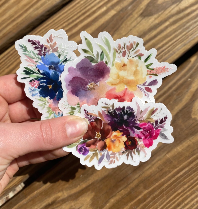 Op de afbeelding: Twee aquarelbloemenstickers met een witte achtergrond. De eerste sticker heeft een boeket met paarse, blauwe en gele bloemen. De tweede sticker heeft een boeket met paarse, rode en gele bloemen.