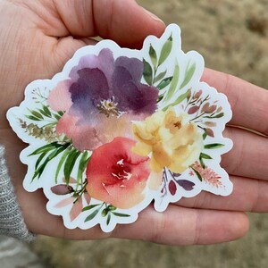 Op de afbeelding: Een aquarel sticker met een boeket bloemen in tinten van paars, geel, roze en groen. De bloemen zijn in een losse, organische stijl gerangschikt, en de sticker heeft een witte achtergrond.