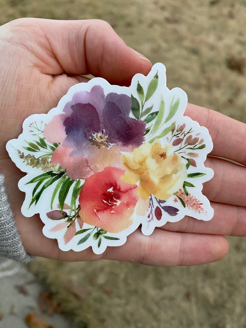 Op de afbeelding: Een aquarel sticker met een boeket bloemen in tinten van paars, geel, roze en groen. De bloemen zijn in een losse, organische stijl gerangschikt, en de sticker heeft een witte achtergrond.