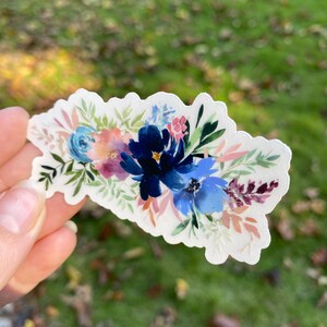 Op de afbeelding: Een sticker met een aquarel bloemenboeket met blauwe, roze en groene bloemen. De bloemen zijn in een cluster gerangschikt en hebben een zachte, romantische uitstraling.