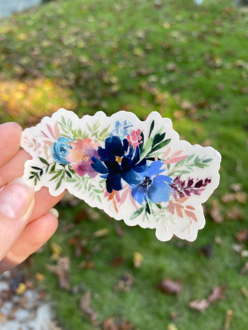 Op de afbeelding: Een sticker met een aquarel bloemenboeket met blauwe, roze en groene bloemen. De bloemen zijn in een cluster gerangschikt en hebben een zachte, romantische uitstraling.
