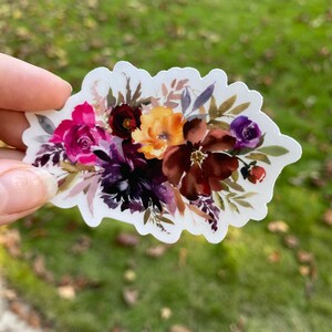 Op de afbeelding: Een aquarel sticker met een boeket van bordeauxrode, roze, paarse en gele bloemen met groene bladeren.