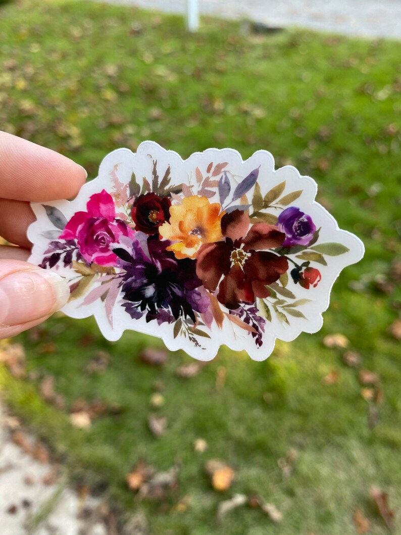 Op de afbeelding: Een aquarel sticker met een boeket van bordeauxrode, roze, paarse en gele bloemen met groene bladeren.