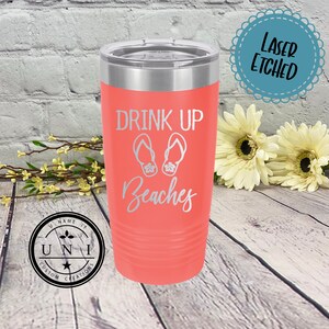 May include: A coral pink stainless steel tumbler with a silver lid. The tumbler has the text "Drink Up Beaches" with a graphic of flip flops. The tumbler is laser etched.