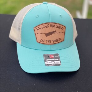 May include: A light blue and white trucker hat with a brown leather patch that says "KILLING MY LIVER ON THE RIVER" with a drawing of a bottle of beer in the water. The hat has a Richardson logo and says "LOW-PRO 115 TRUCKER".
