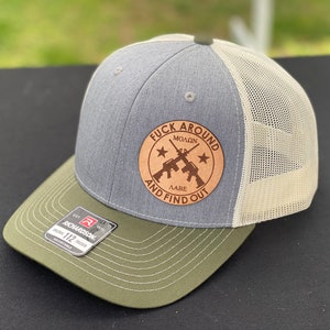 May include: A gray and green trucker hat with a brown leather patch that says "F**k Around And Find Out" with a crossed rifle design and two stars.