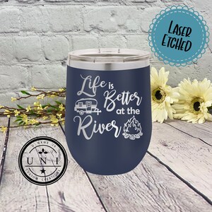 May include: A navy blue stainless steel tumbler with a white design that says "Life is Better at the River" with a camper and campfire graphic. The tumbler is laser etched.