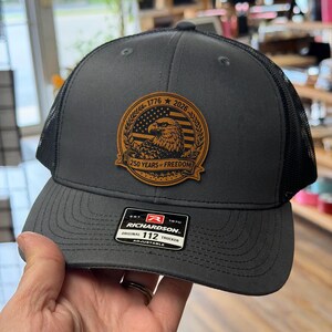 May include: A charcoal gray trucker hat with a black mesh back and a brown leather patch on the front. The patch features an eagle, an American flag, and the text 1776 2026 250 YEARS OF FREEDOM. A black sticker on the brim reads RICHARDSON 112.
