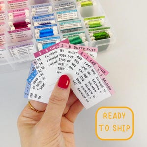 May include: A hand holds color cards for embroidery floss, displaying shades of pink, blue, green, and purple. The cards list color names and numbers. A clear plastic organizer filled with floss is in the background. Text on the cards includes "Fuchsia" and "Dusty Rose".
