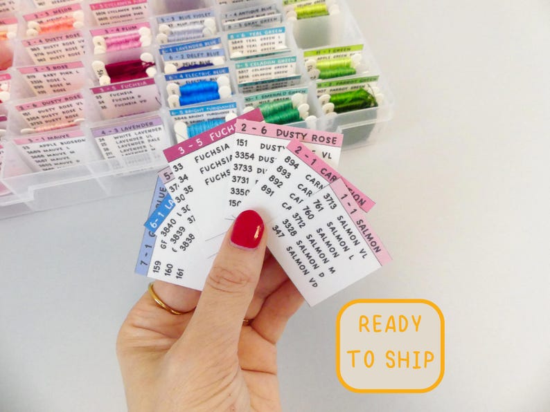 May include: A hand holds color cards for embroidery floss, displaying shades of pink, blue, green, and purple. The cards list color names and numbers. A clear plastic organizer filled with floss is in the background. Text on the cards includes "Fuchsia" and "Dusty Rose".