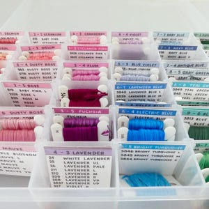 May include: A clear plastic organizer filled with spools of colorful embroidery floss. Each spool is labeled with a color name and number, including shades of pink, blue, green, and purple. The floss is neatly arranged for easy access.