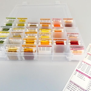May include: A clear plastic organizer filled with spools of colorful embroidery floss. The floss is arranged by color in small compartments, with labels indicating the color names and numbers. A color chart is visible on the right.