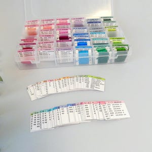 May include: A clear plastic organizer filled with spools of colorful embroidery floss. The floss is arranged by color, with labels indicating the color names and numbers. Several color-coded cards are also visible.