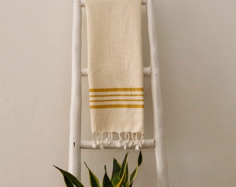 May include: A cream-colored towel with mustard yellow stripes and fringe hangs on a white wooden ladder. The towel is made of a textured fabric. A green and yellow plant is in the foreground.