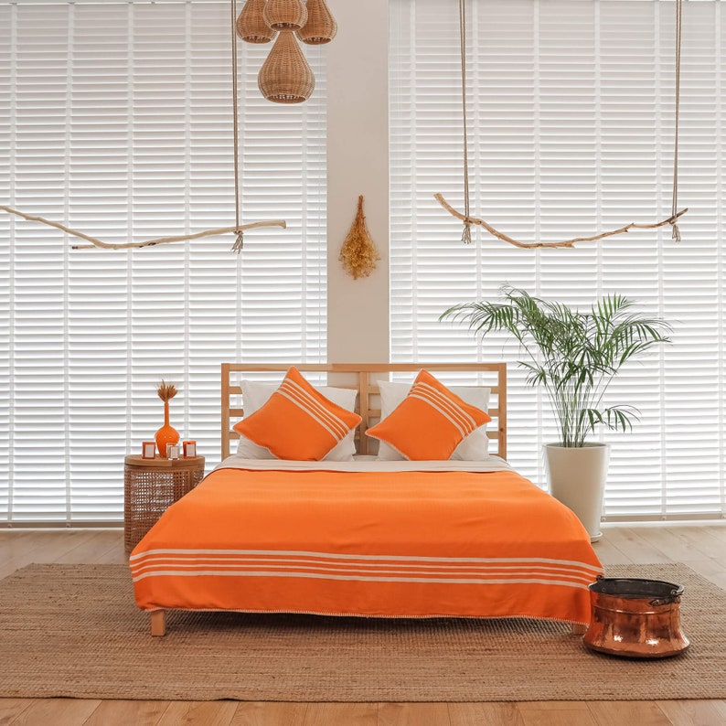 May include: A bed with a bright orange striped duvet cover and two white and orange pillows. The bed is made of wood and has a headboard. The bed is in a room with white blinds and a potted plant.