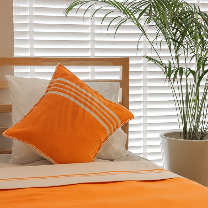 May include: A white bed with an orange pillow with white stripes. The pillow has a white border. The bed is made with a white sheet and a white and orange blanket. A green plant is in a white pot in the background.