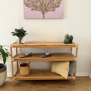 Puede incluir: Una estantería de bambú de tres niveles con un acabado natural. La estantería tiene tres estantes y está hecha de bambú tejido. La estantería está decorada con un jarrón de bayas rojas, dos pequeñas tazas de cerámica azul y blanca y dos pequeños cactus verdes.