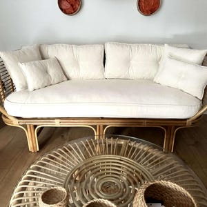 May include: A white cushioned rattan sofa with a woven frame and a round woven coffee table with a glass top. The sofa has a white cushion and several white pillows.