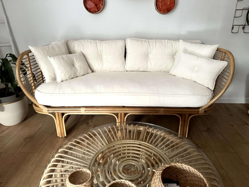 May include: A white cushioned rattan sofa with a woven frame and a round woven coffee table with a glass top. The sofa has a white cushion and several white pillows.