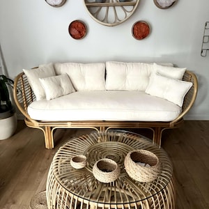 May include: A white wicker sofa with white cushions and a round wicker coffee table with three woven baskets on top. The sofa and table are in a room with white walls and a wooden floor.