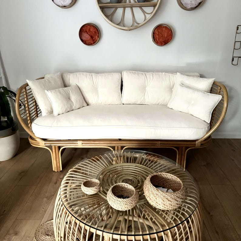 May include: A white wicker sofa with white cushions and a round wicker coffee table with three woven baskets on top. The sofa and table are in a room with white walls and a wooden floor.