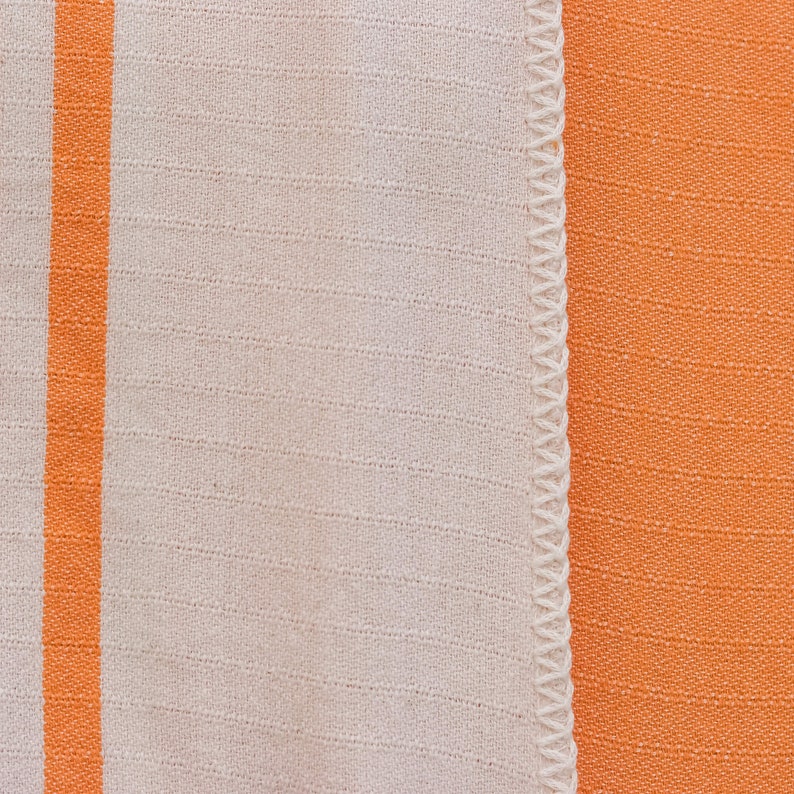 May include: A close-up of a white fabric with a single orange stripe running vertically down the left side. The fabric is sewn to a solid orange fabric.