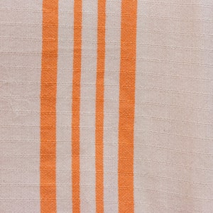 May include: A close-up of a white fabric with orange stripes. The fabric has a woven texture.