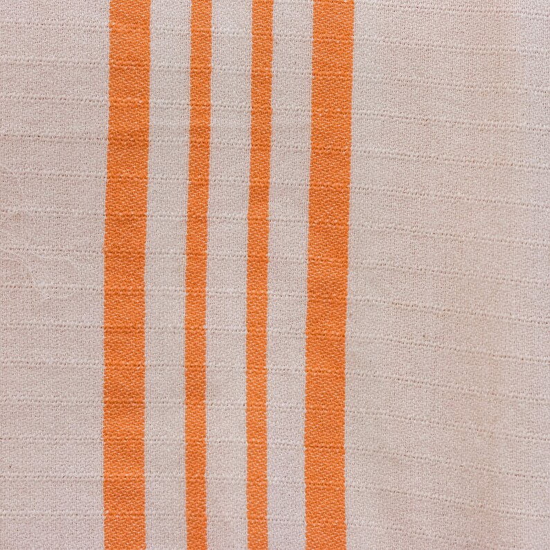 May include: A close-up of a white fabric with orange stripes. The fabric has a woven texture.