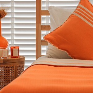 May include: A close-up of a bed with orange bedding and a white and orange pillow. The bed is made with a white sheet and a white and orange striped pillow. The bed is next to a small wooden table with two candles on it.