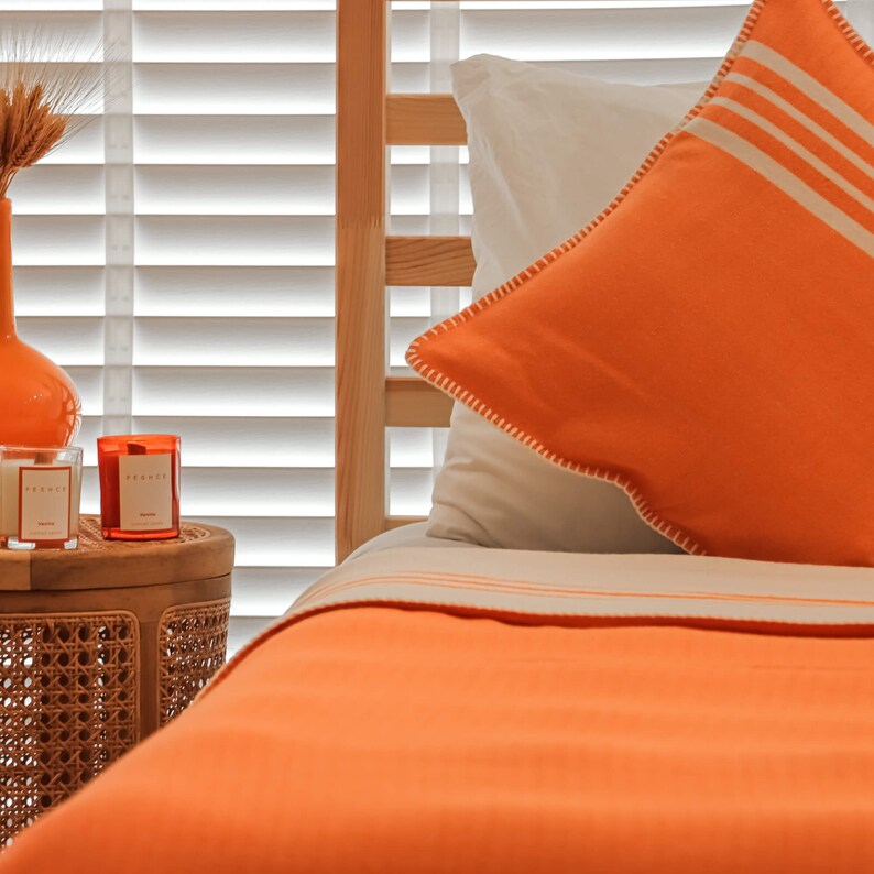 May include: A close-up of a bed with orange bedding and a white and orange pillow. The bed is made with a white sheet and a white and orange striped pillow. The bed is next to a small wooden table with two candles on it.